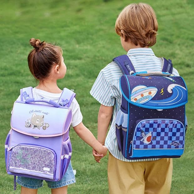 Two school-age children walking away hand in hand across grass wearing patterned Spinecare backpacks